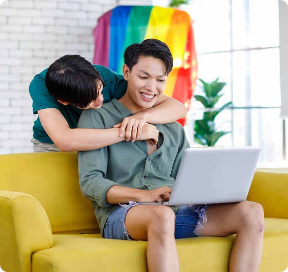 a woman hugs a young man on a couch - a pride flag hangs in the background